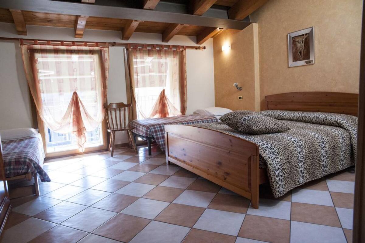 A cozy room with a wooden bed, a chair, and a small table. There are two windows with curtains, and a picture on the wall.
