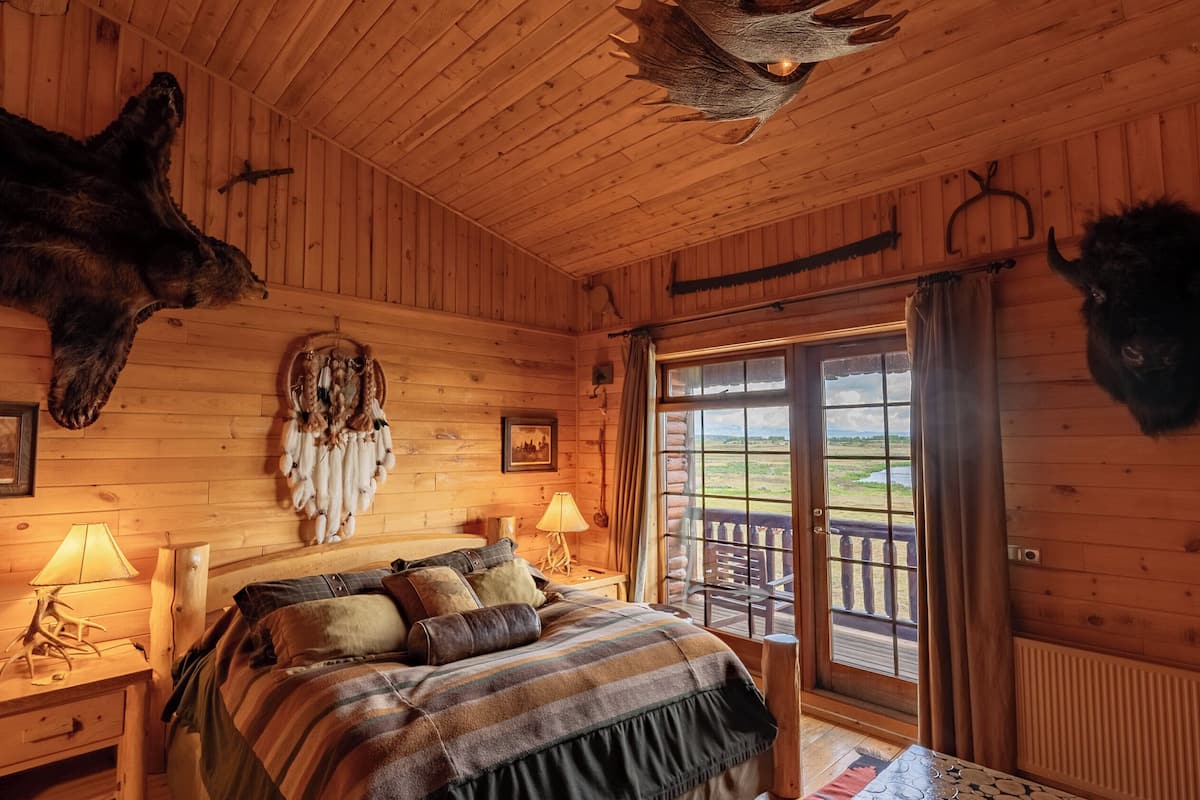 Quarto de cabana de tronco com uma cama, duas luminárias, um apanhador de sonhos e cabeças de animais montadas na parede.