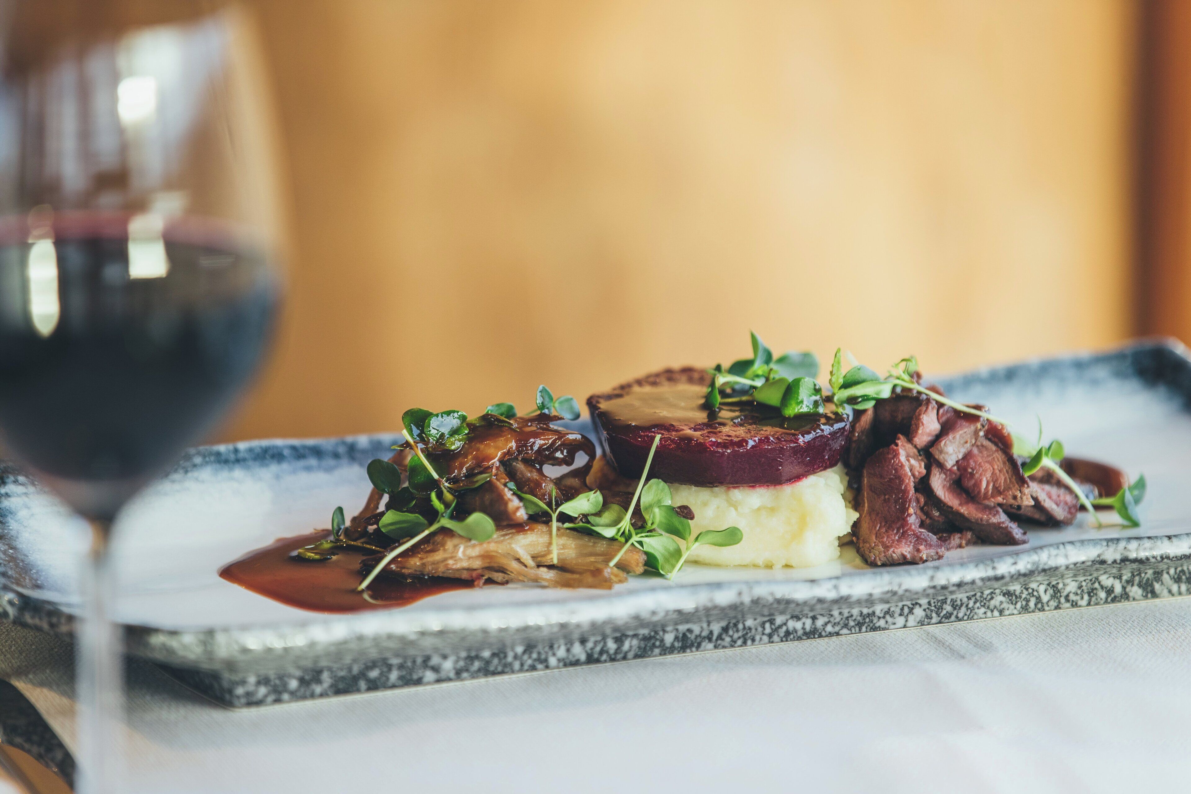 A plated dish with meat, mushrooms, and greens, accompanied by a glass of red wine.