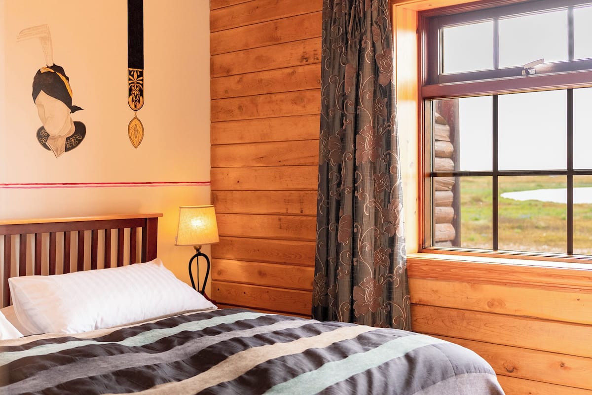 A wooden cabin room with a bed, a lamp, and a window with curtains.