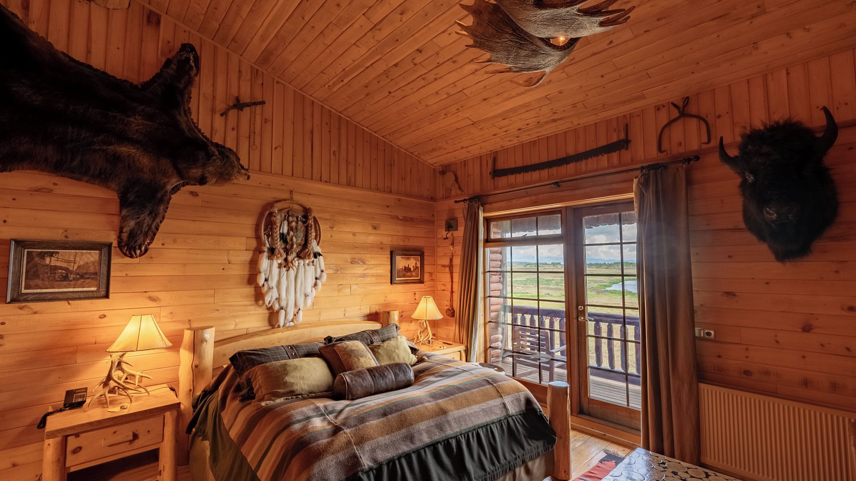 Une chambre dans un chalet en rondins, avec un lit, deux lampes, un attrape-rêves et des têtes d’animaux montées au mur.