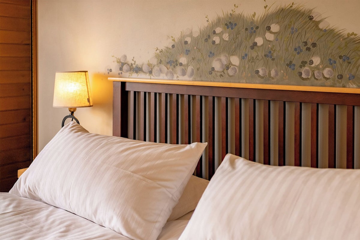 Quarto de hotel com cama, duas almofadas, cabeceira com ripas verticais e luminária fixada na parede.