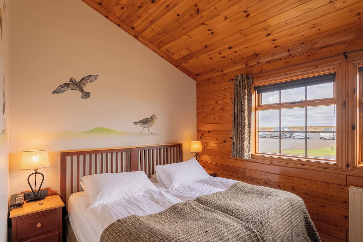 Quarto de cabana de madeira com uma cama, duas luminárias de cabeceira, uma mesa de cabeceira e uma janela com vista para um estacionamento.