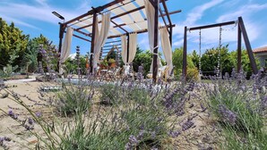 Una estructura de pérgola de madera con cortinas de tela blanca, rodeada de plantas de lavanda y mobiliario de exterior.