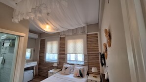 Un dormitorio con panel de pared de madera, dos ventanas con cortinas estampadas, una cama con ropa blanca, una mesita de noche con lámpara y un techo decorado con tela.