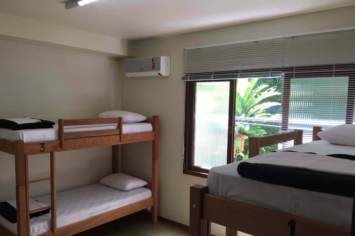 A dormitory room with bunk beds, a window with blinds, and an air conditioning unit.