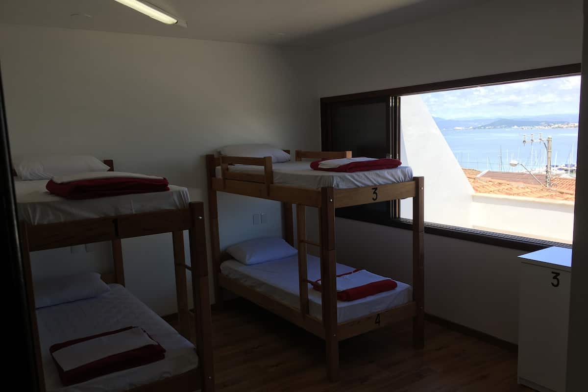 A dormitory room with bunk beds numbered 2, 3, and 4, a window with a view of the sea, and a small desk with a chair.