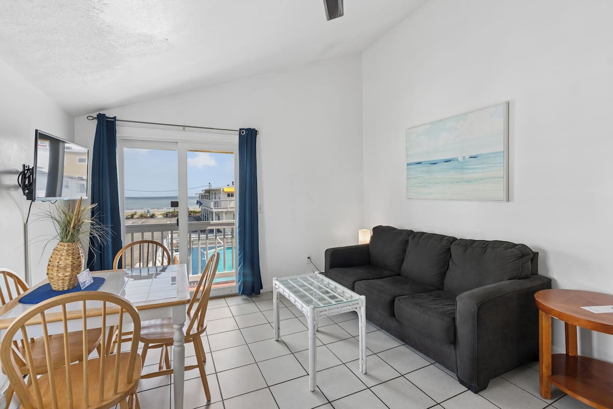 A living room with a sofa, a small table, a dining table with chairs, a TV, and a view of the sea through a balcony.