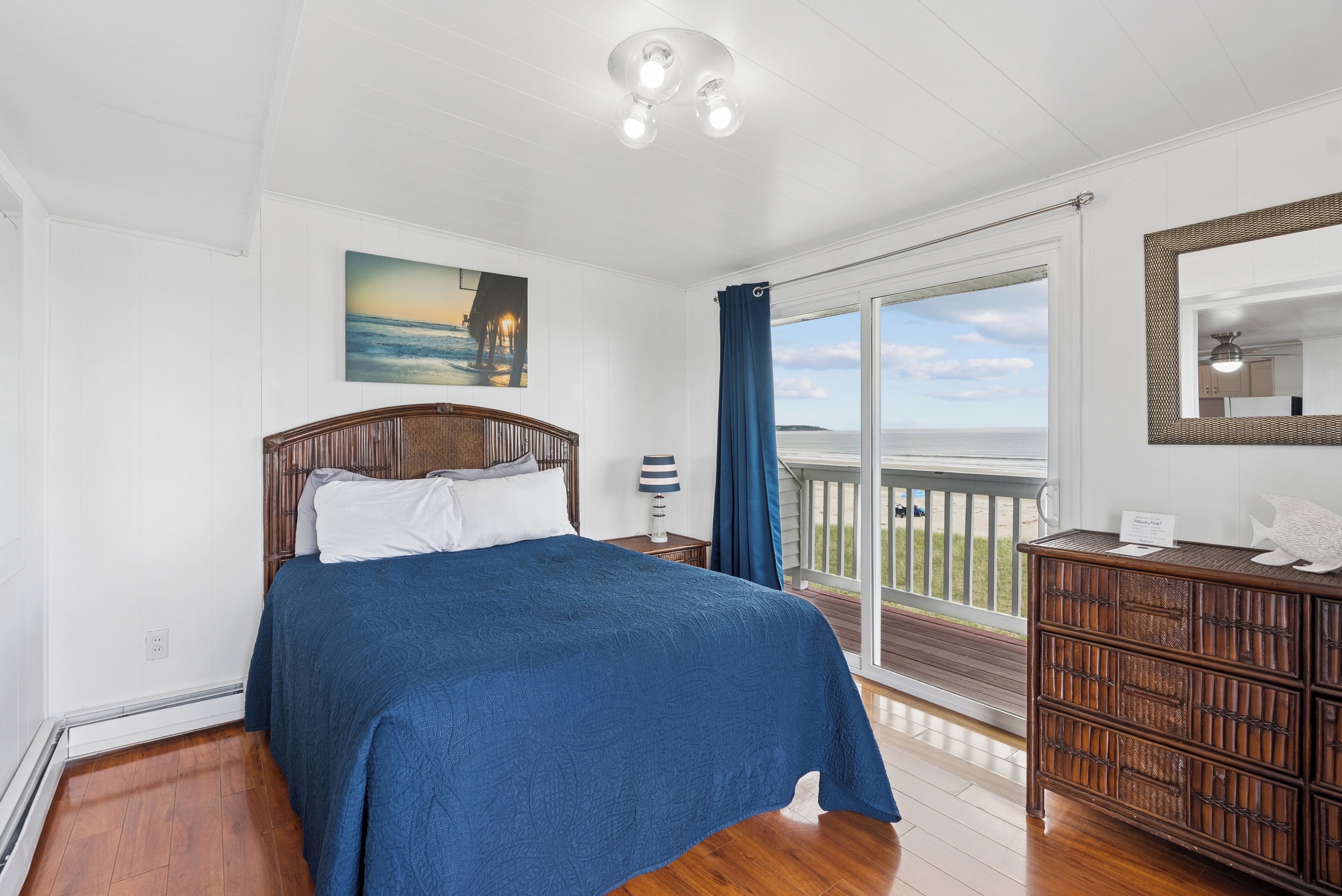 A bedroom with a bed, a dresser, a painting, and a view of the ocean.