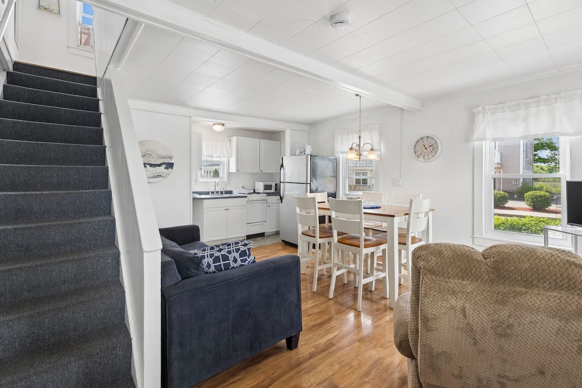 A modern kitchen with white cabinets, a dining area with a wooden table and chairs, a gray sofa with a patterned pillow, and a staircase leading to another level.