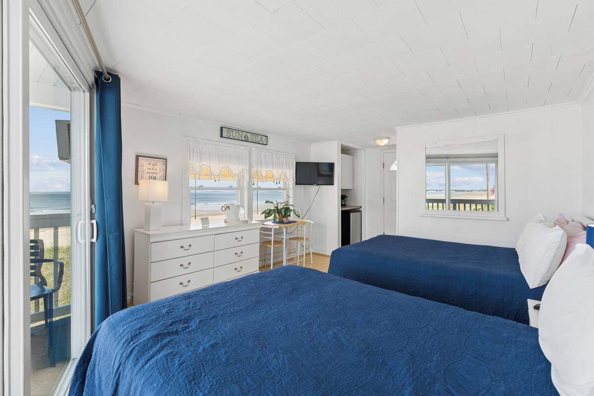 A bedroom with two beds, a desk, a chair, a TV, and a view of the sea.