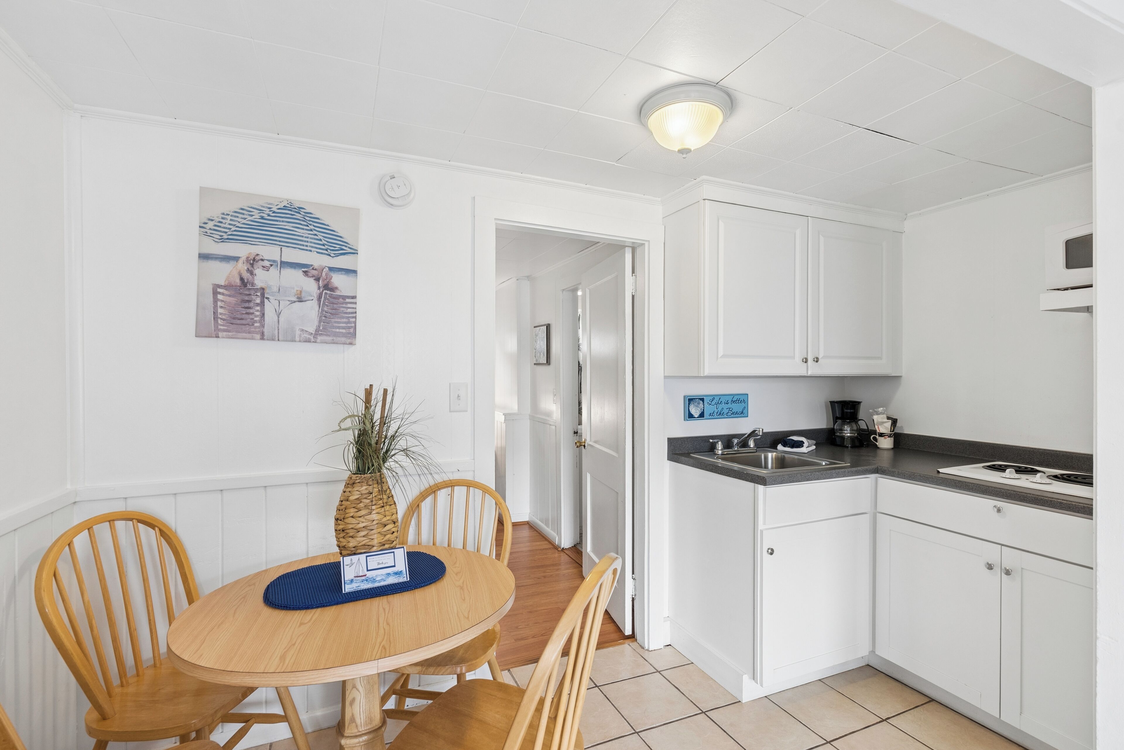 A kitchen with white cabinets, a wooden dining table with chairs, a pineapple centerpiece, and a painting on the wall.