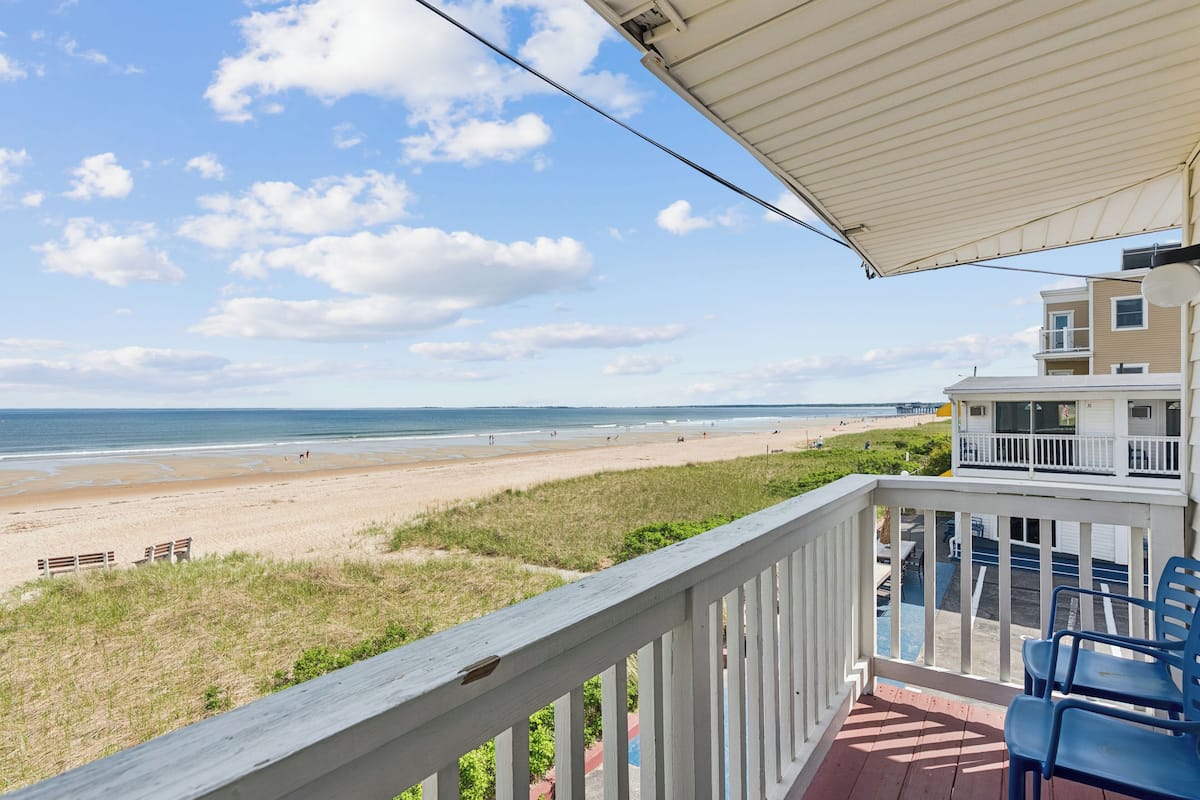 A balcony with a view of a beach, a covered area, and a building with a balcony.