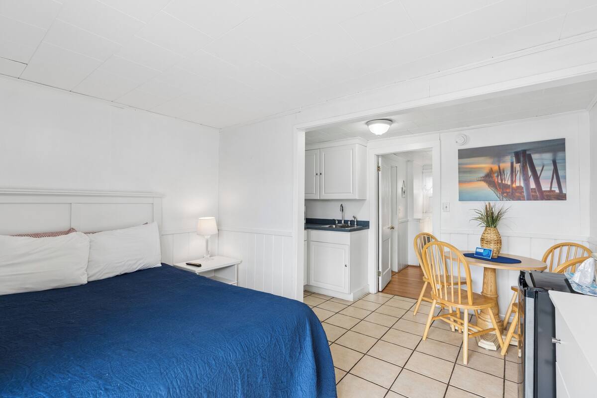 A hotel room with a bed, a small desk, a chair, a table, a kitchen area, and a painting on the wall.