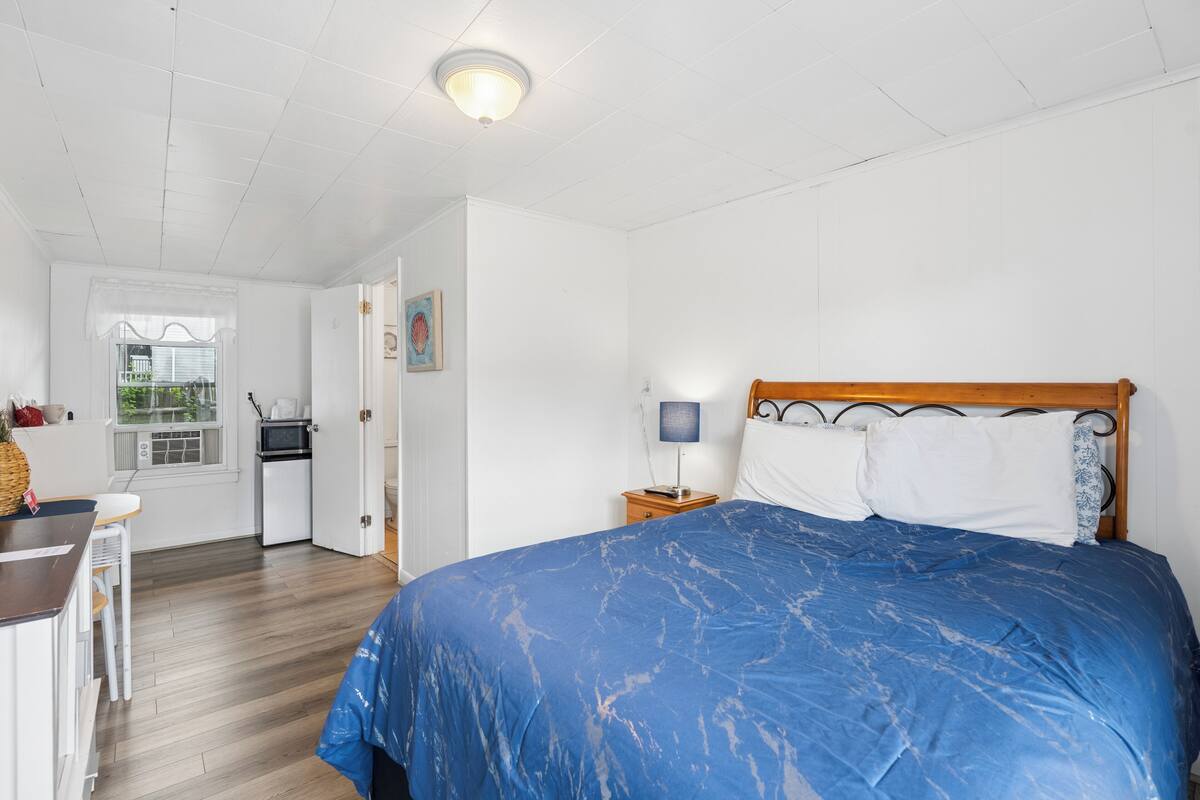 A bedroom with a bed, a nightstand, a lamp, a window, and a kitchenette.