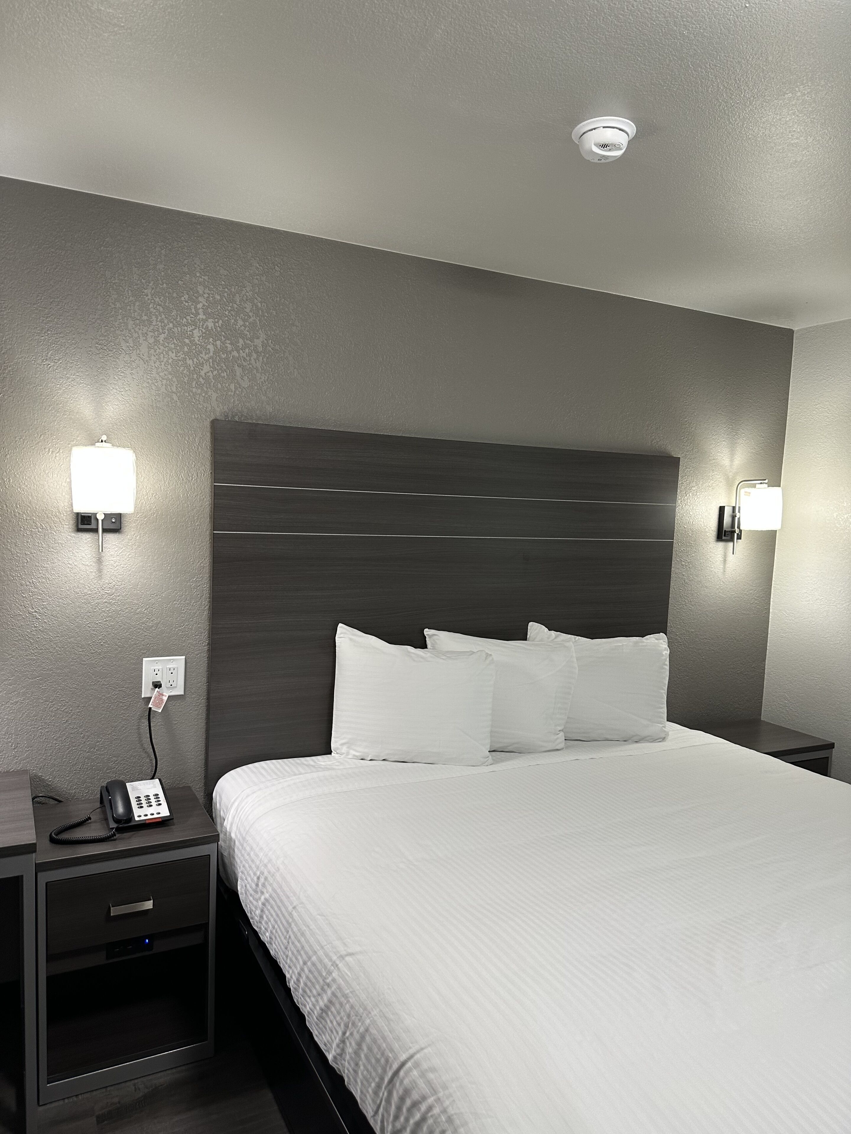Una habitación de hotel con una cama grande, dos mesitas de noche, un teléfono e iluminación en la pared.