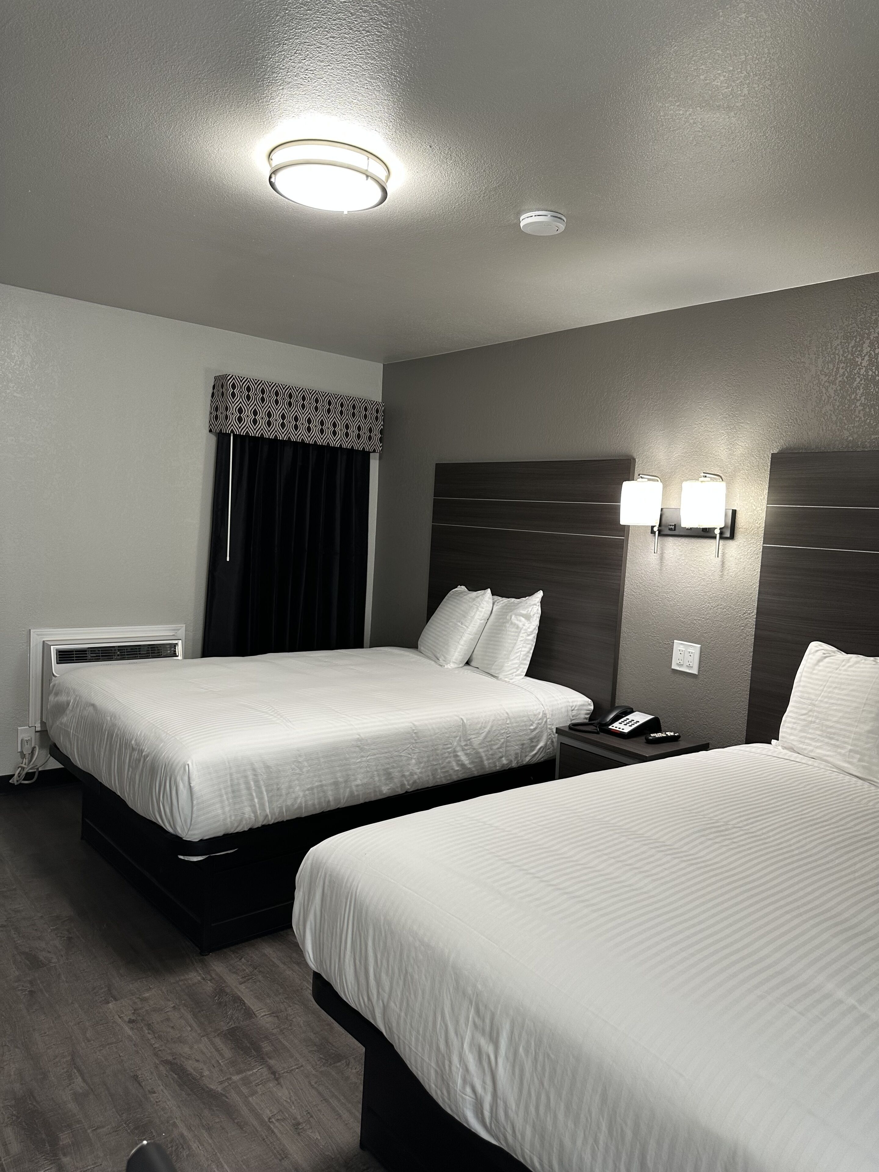 A hotel room with two beds, a bedside table with a phone, and a wall-mounted light fixture.