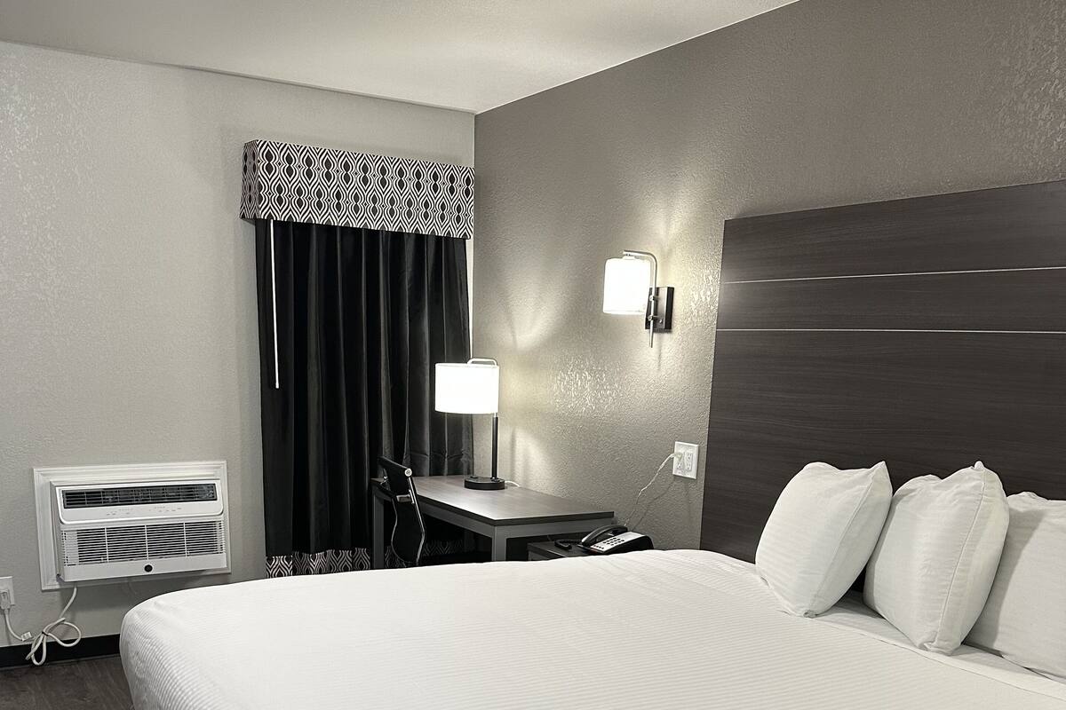A hotel room with a large bed, a desk, a chair, a lamp, a phone, a wall-mounted light, a curtain, and an air conditioning unit.