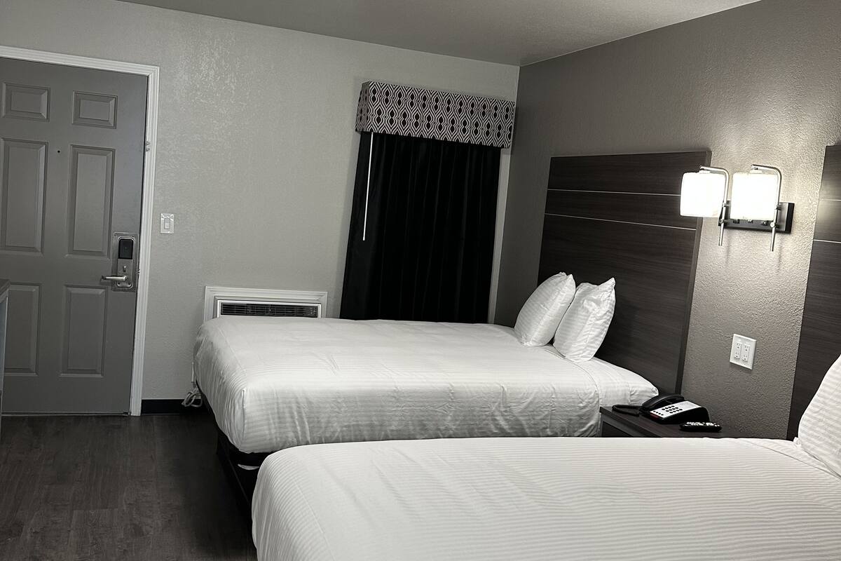 A hotel room with two beds, a bedside table with a phone and remote, a wall-mounted light, and a door with a keypad lock.