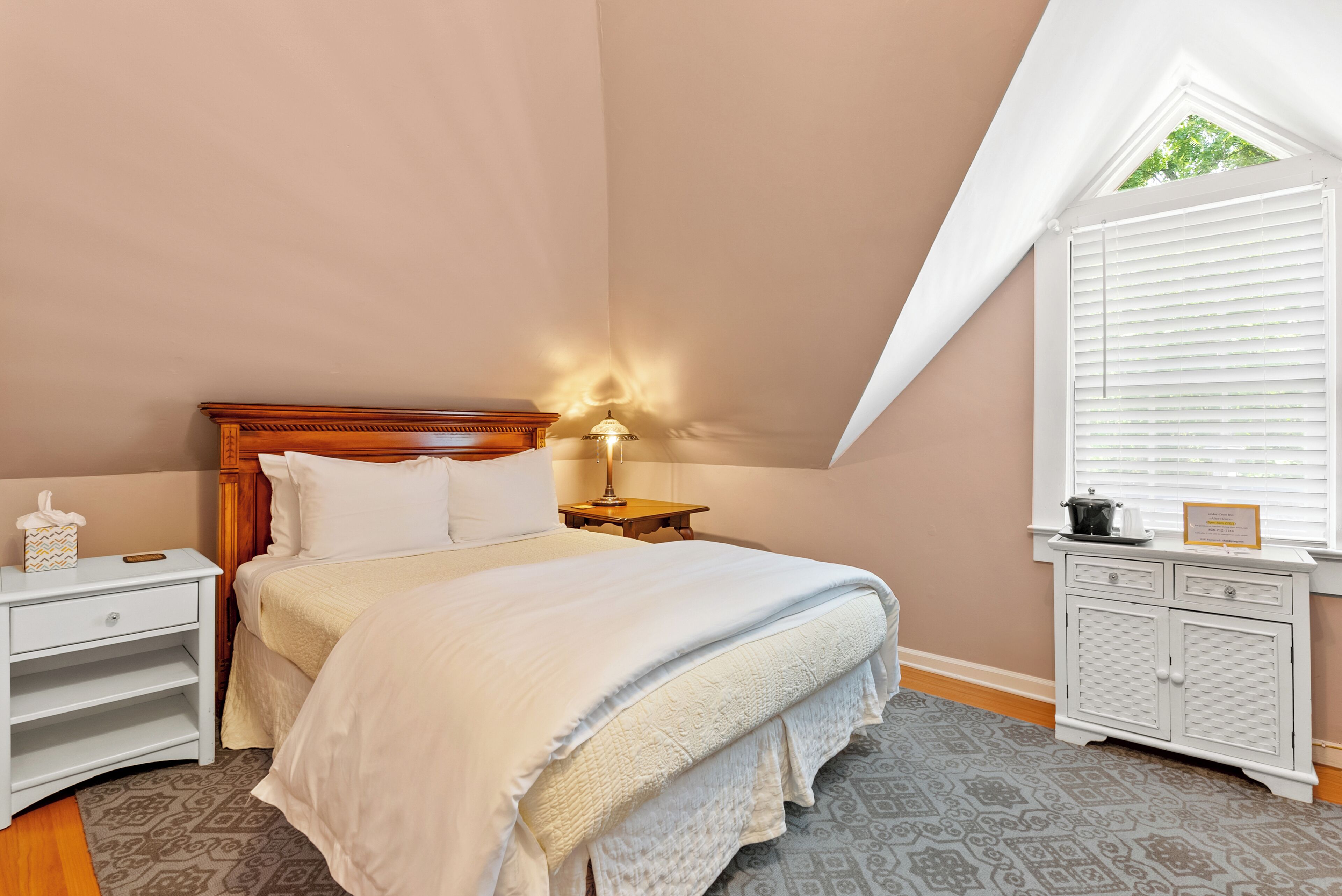 A bedroom with a wooden headboard, a white bedspread, a bedside table with a lamp, a nightstand, and a window with blinds.