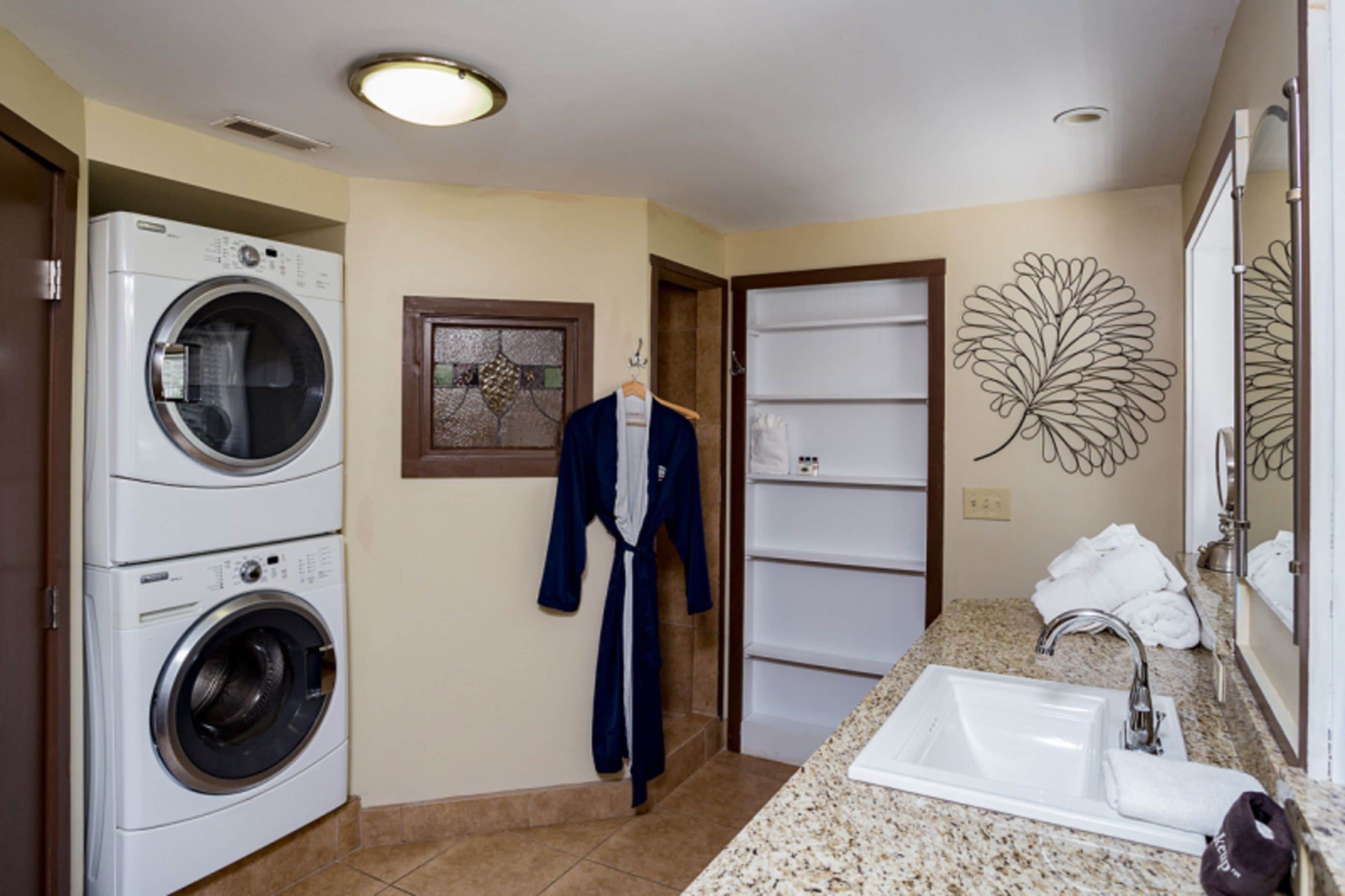 A hotel bathroom with a washing machine, a robe hanging on the wall, a sink, and a decorative leaf wall art.