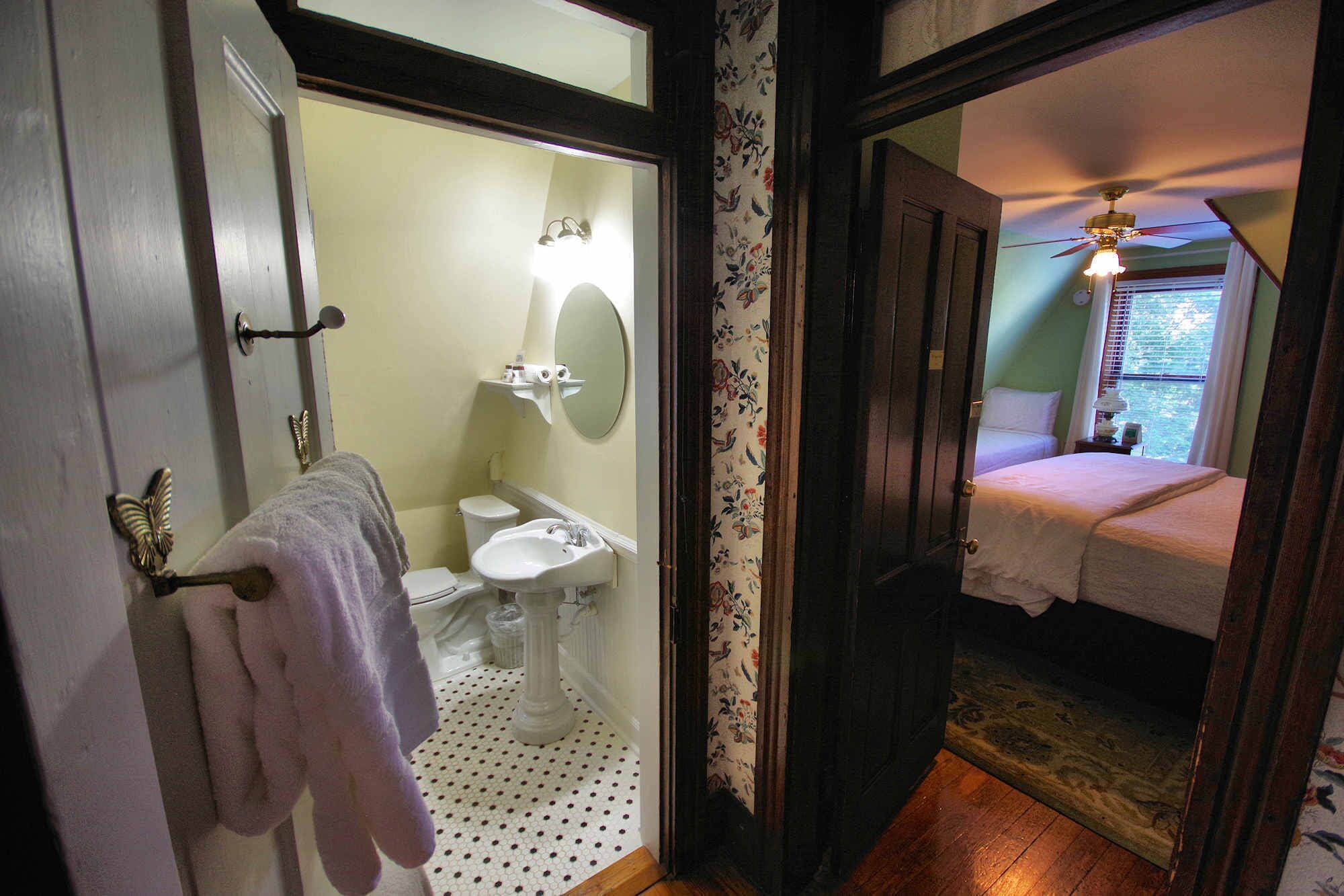 Une salle de bain avec des toilettes, un lavabo et un miroir. La pièce adjacente contient un lit et un ventilateur de plafond.