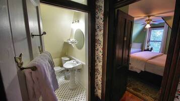 Une salle de bain avec des toilettes, un lavabo et un miroir. La pièce adjacente contient un lit et un ventilateur de plafond.