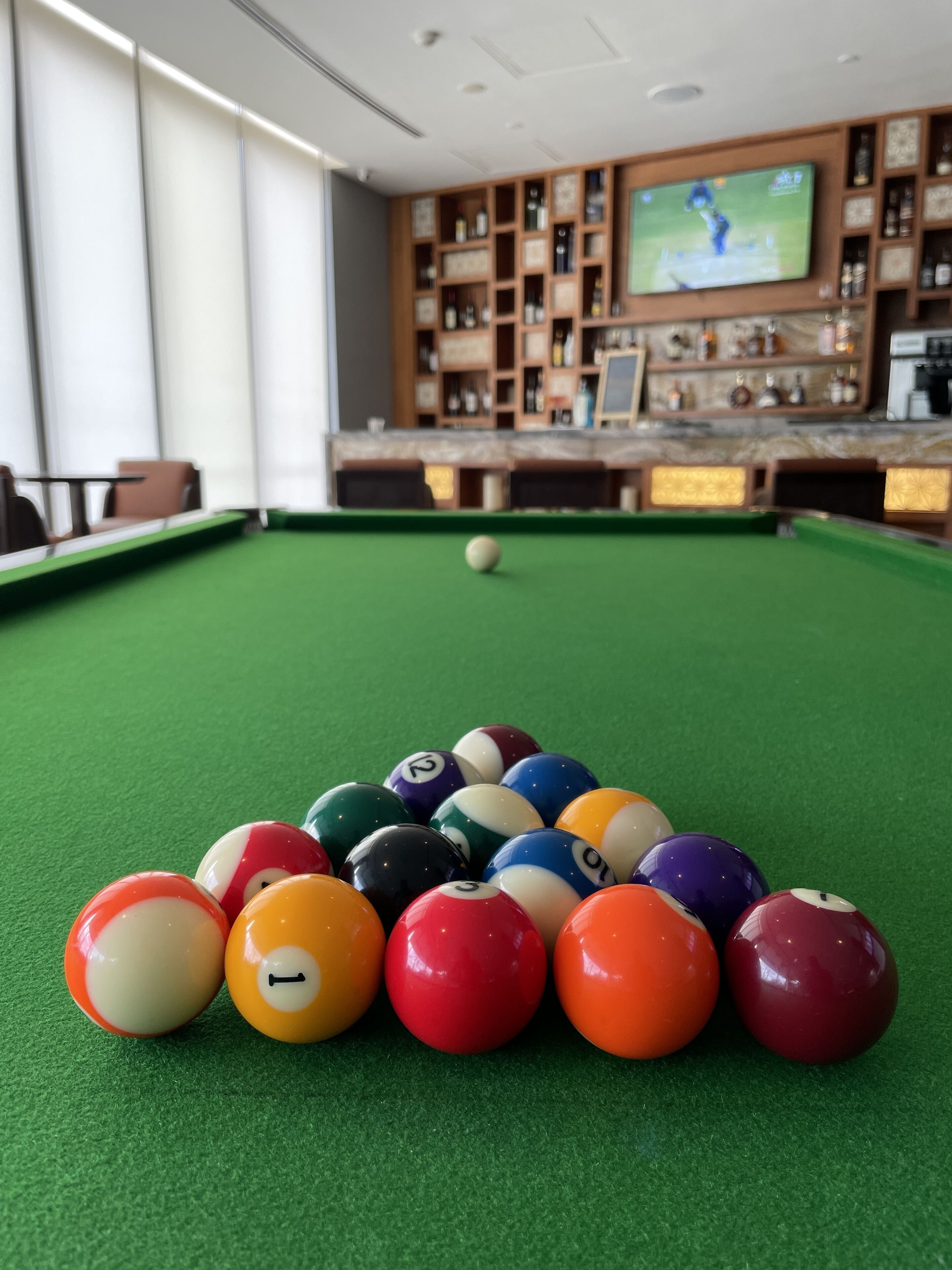A pool table with a variety of colored balls, a television displaying a sports event, and a bar area with shelves of bottles and glasses.