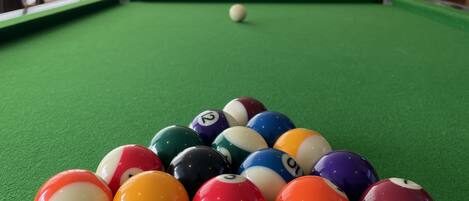 A pool table with a variety of colored balls, a television displaying a sports event, and a bar area with shelves of bottles and glasses.