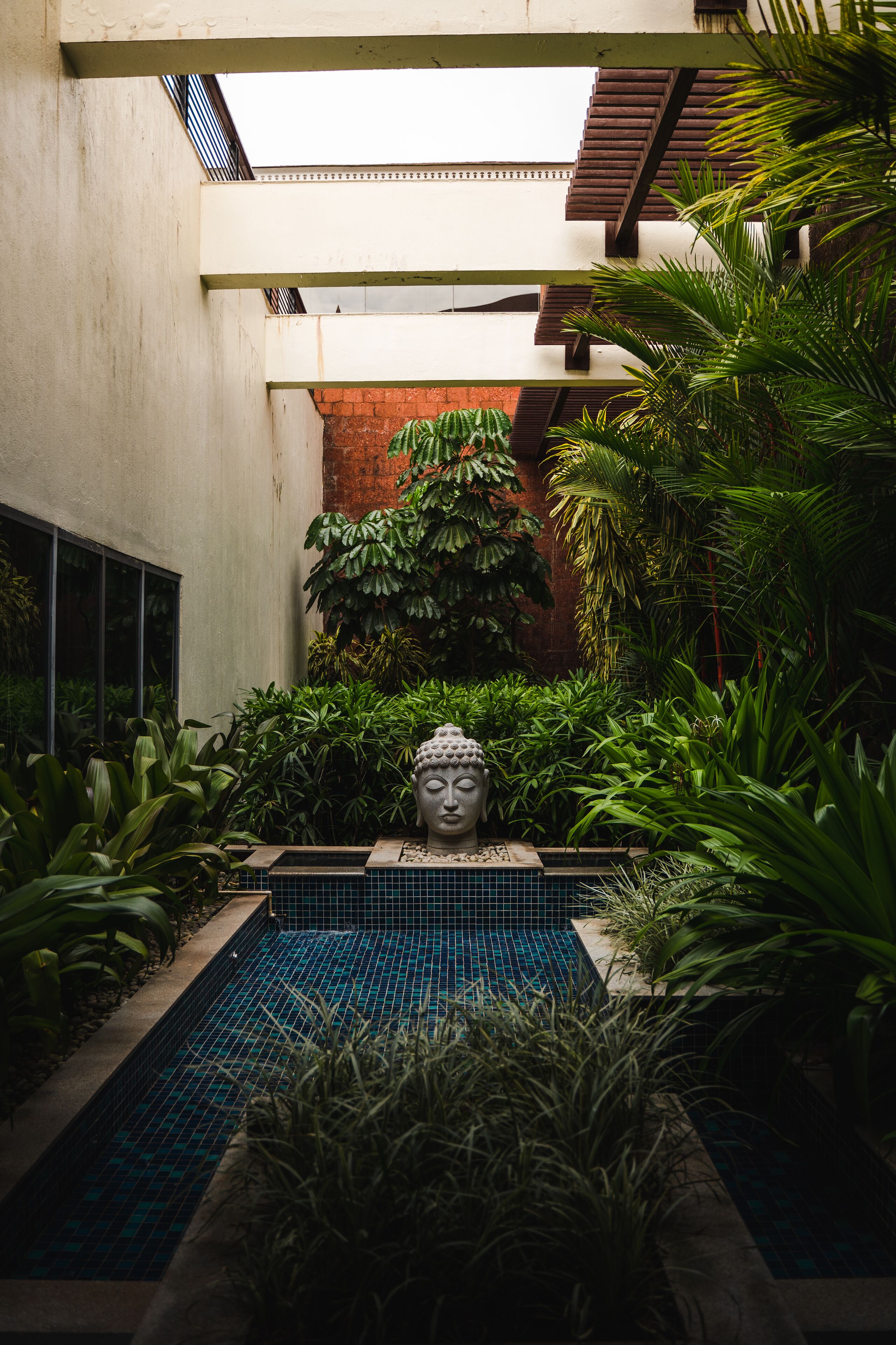 Un jardin intérieur paisible, avec une statue de Bouddha au centre, un petit bassin et une végétation luxuriante.