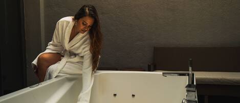A person in a white bathrobe leaning over a bathtub filled with water and orange flower petals.