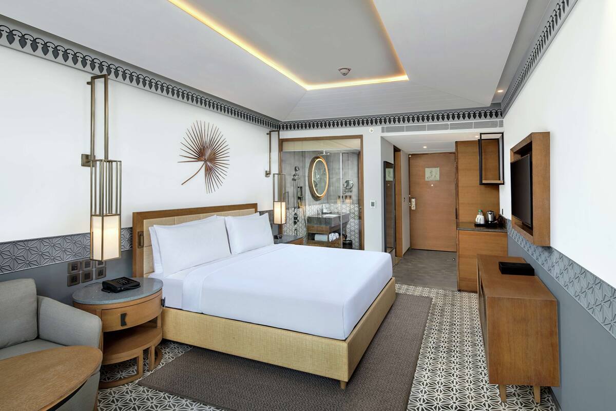 Habitación de hotel con una cama grande, mesitas de noche, televisor y baño visible a través de una puerta abierta.