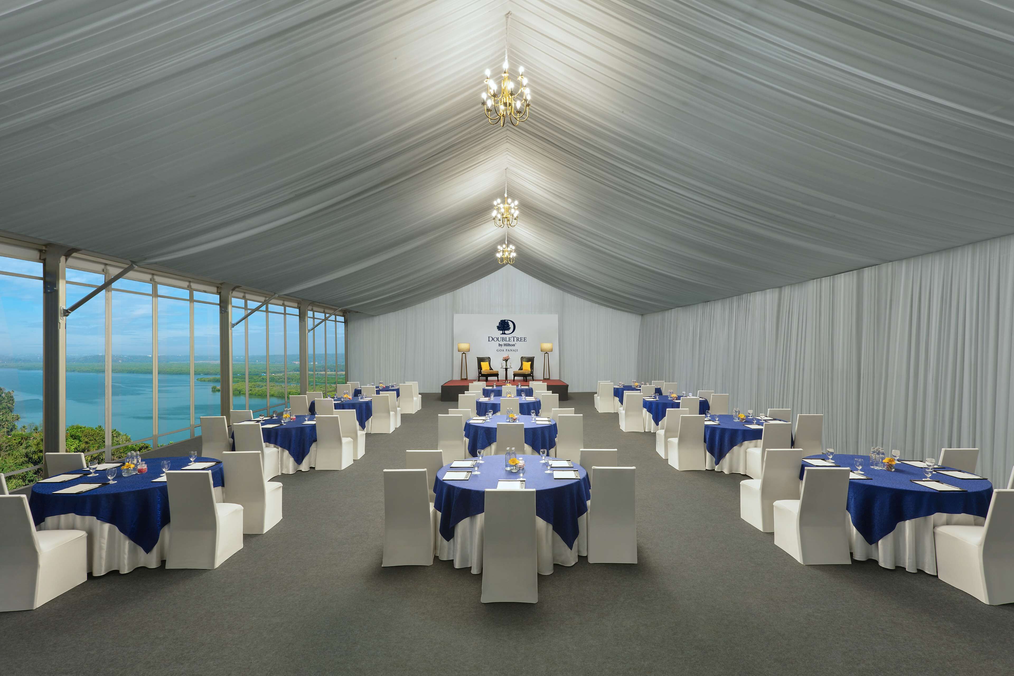 Un salón de banquetes con mesas redondas, sillas blancas y un escenario con un atril.