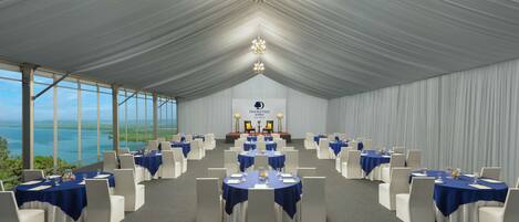 Un salón de banquetes con mesas redondas, sillas blancas y un escenario con un atril.