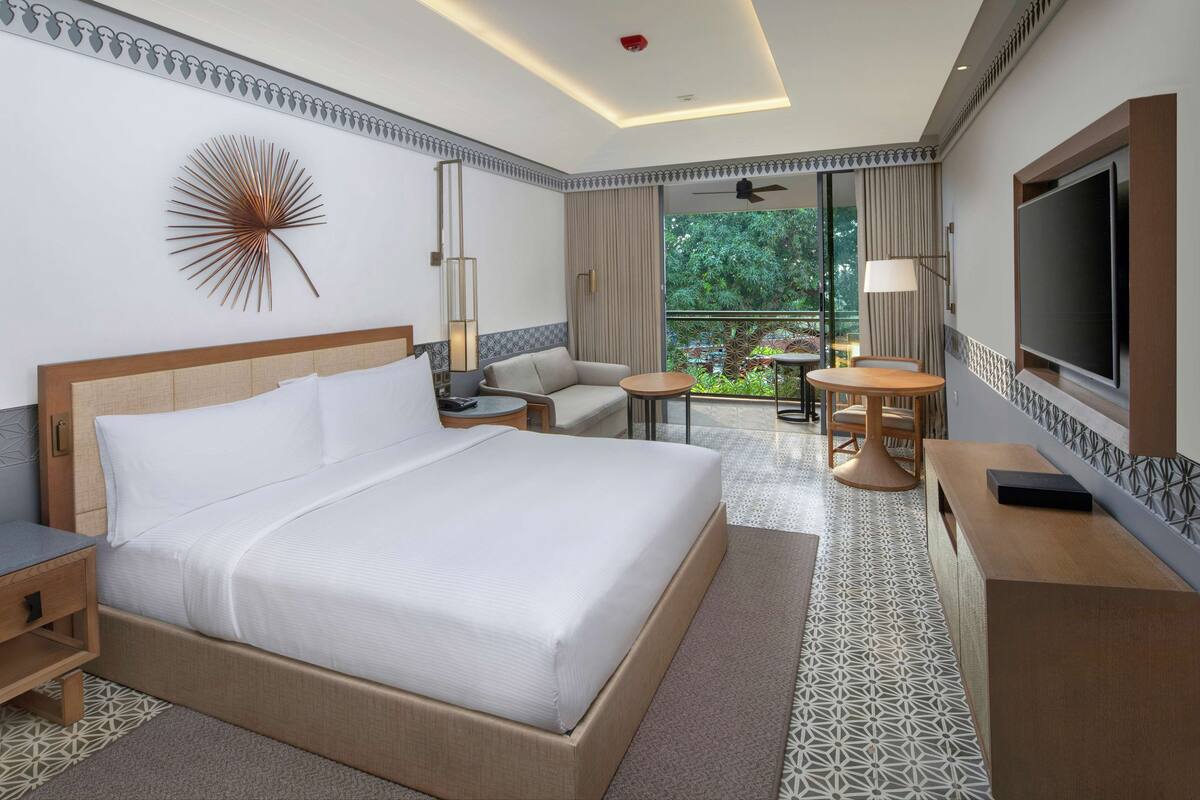 Habitación de hotel con una cama grande, un escritorio, una silla, un televisor y un balcón con vistas a zonas verdes.