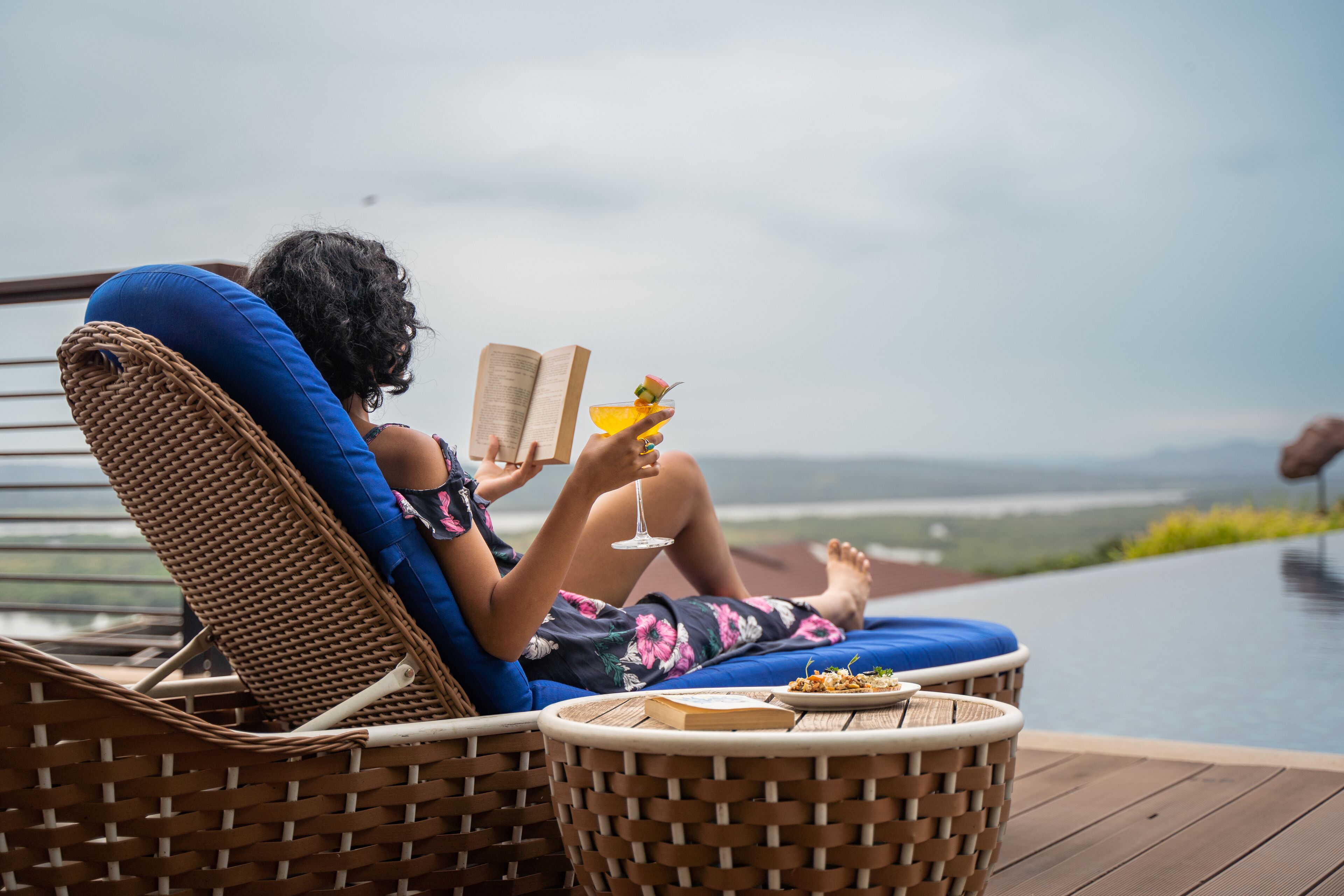 A person relaxing on a wicker lounge chair with a book and a drink, overlooking a pool and distant landscape.