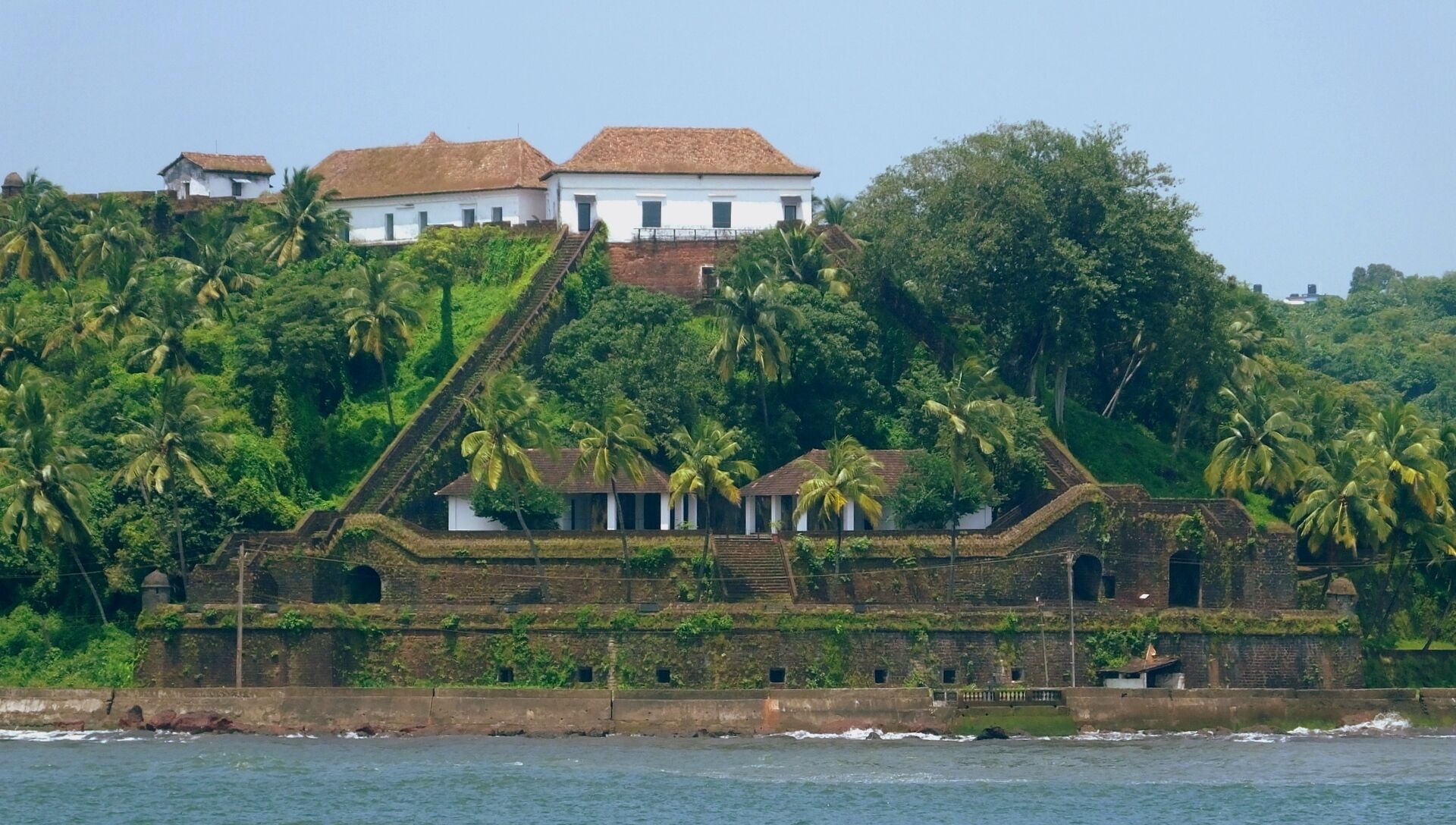 Un edificio costero con una escalera que lleva a una estructura blanca de techo rojo, rodeado de abundante vegetación y palmeras.