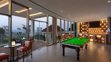 A pool table in a well-lit room with large windows, a bar area with a marble countertop, and seating arrangements.