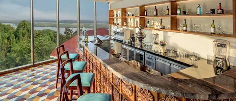 A high-rise bar with a panoramic view of a lake and forest, featuring a patterned floor, bar stools, and a well-stocked shelf with various bottles and glasses.