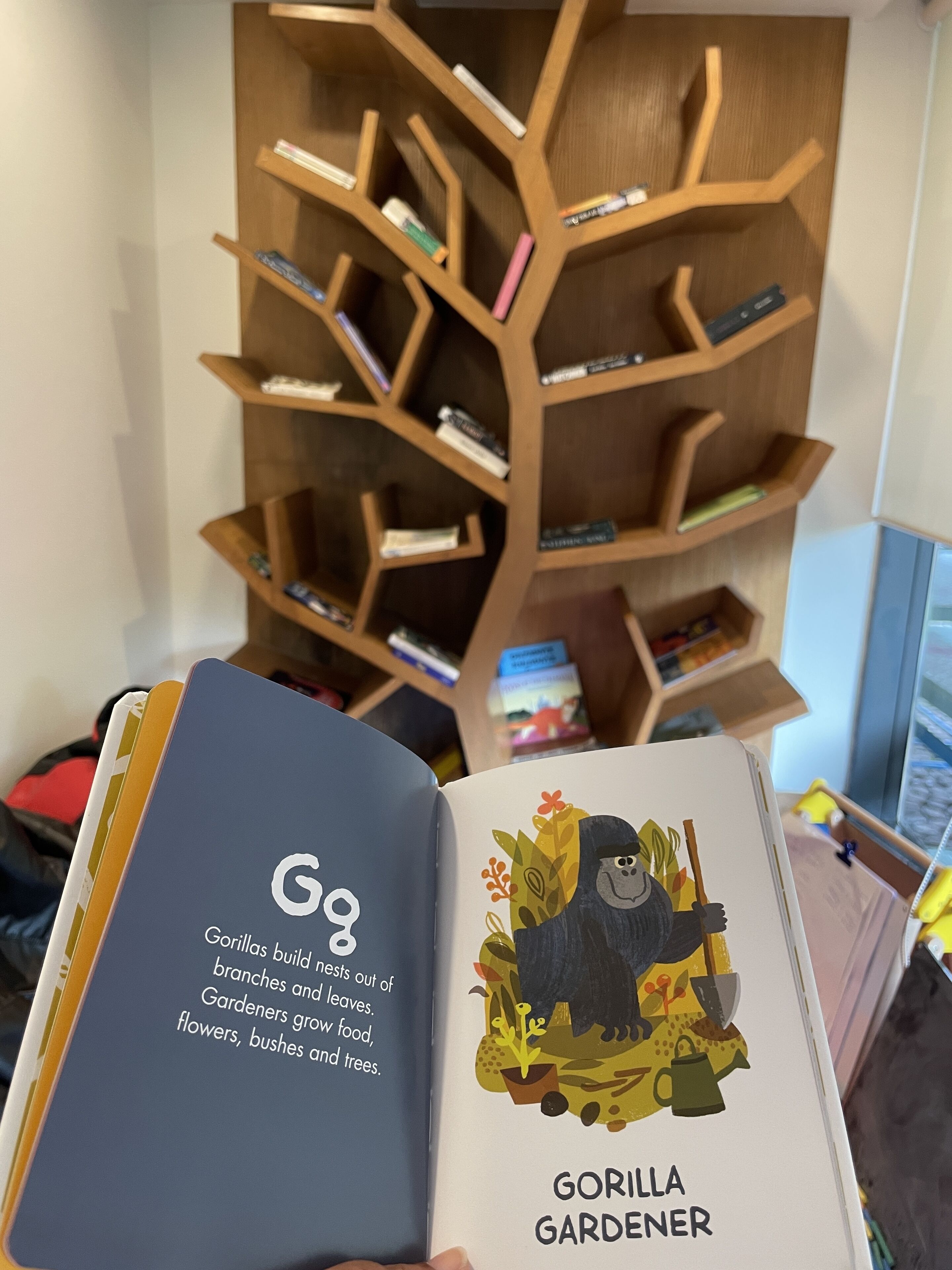 Ein Buch mit dem Titel 'Gorilla Gardener', dessen Cover eine Illustration eines Gorillas inmitten von Pflanzen zeigt. Im Hintergrund ist ein Bücherregal aus Holz mit verschiedenen Büchern und Gegenständen zu sehen.