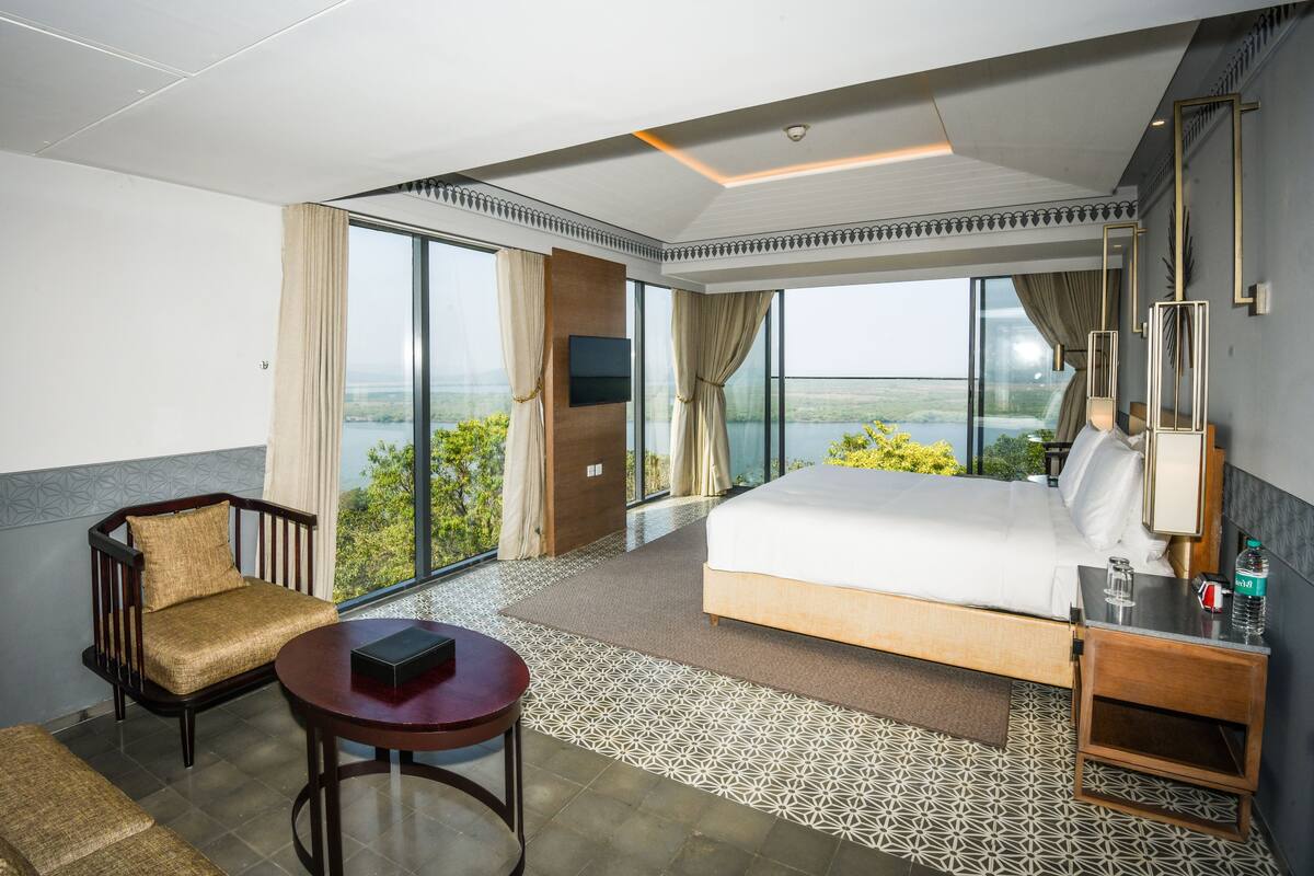 Habitación de hotel con una cama grande, zona de estar con mesa, televisor y vistas al exterior.