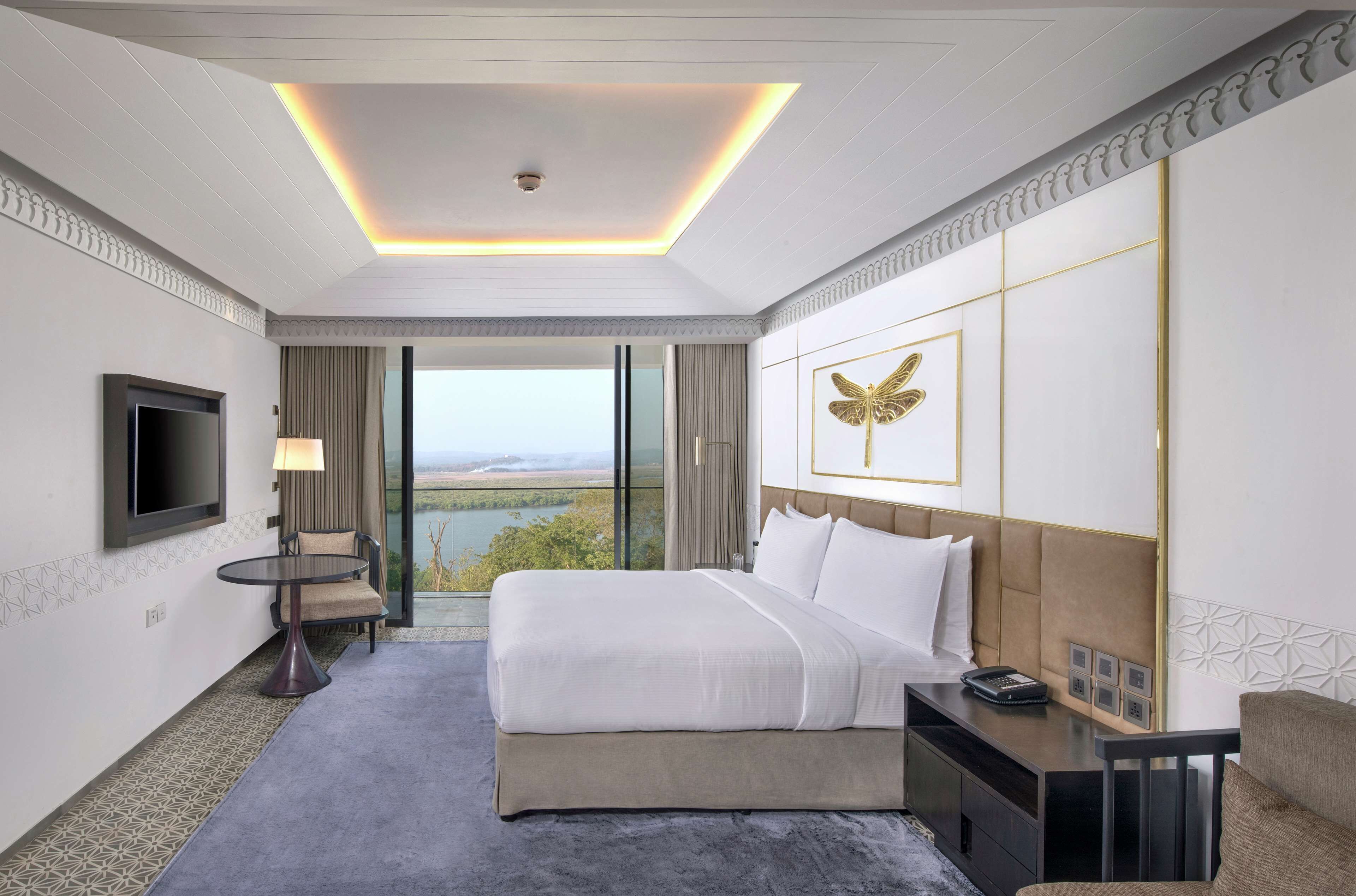 A hotel room with a large bed, a flat-screen TV, a desk with a lamp, and a view of a lake through the window.