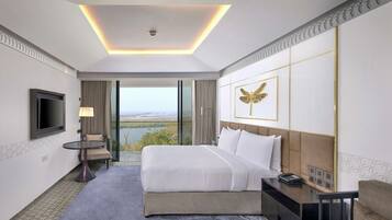 A hotel room with a large bed, a flat-screen TV, a desk with a lamp, and a view of a lake through the window.