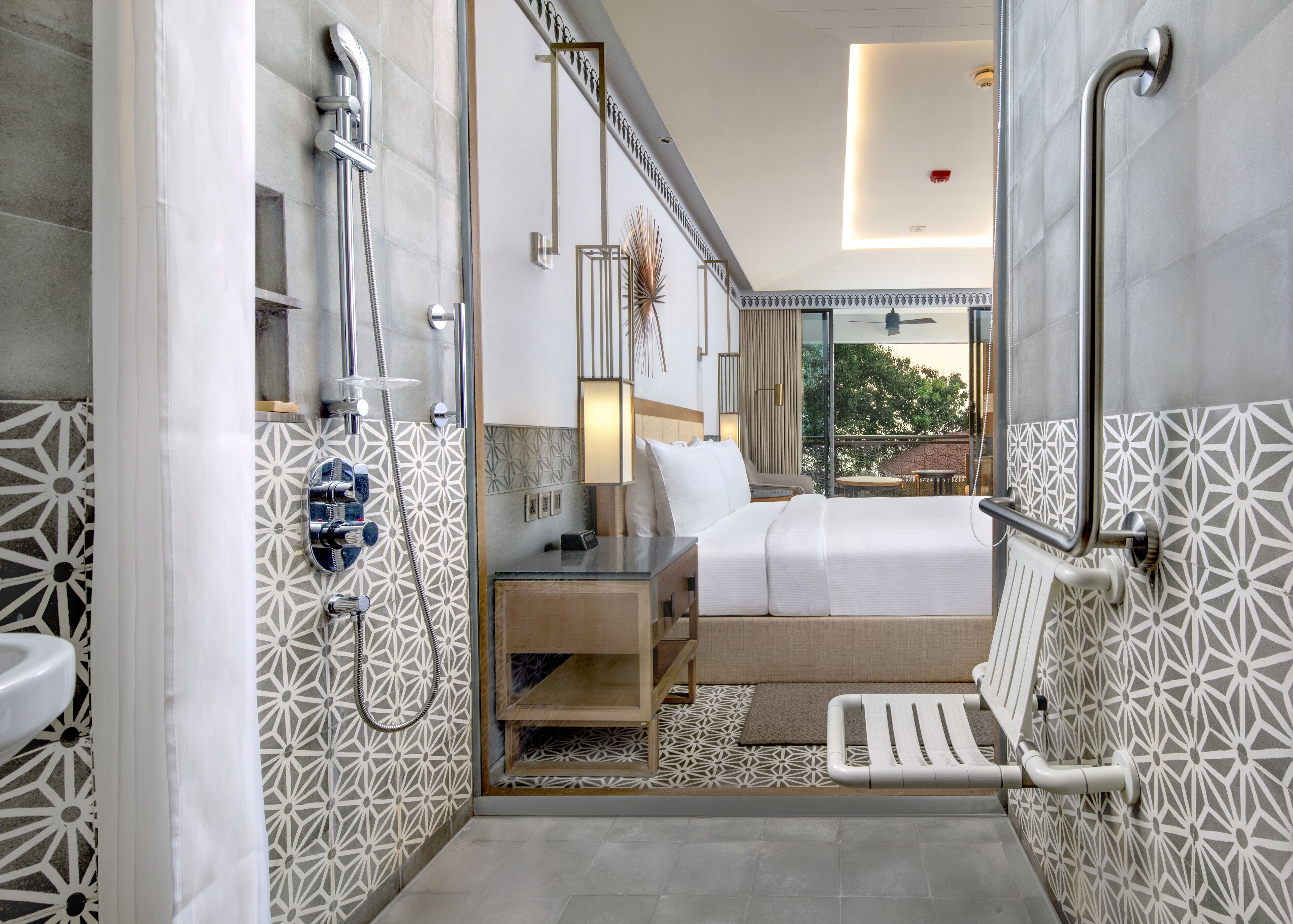 Un baño moderno con ducha, inodoro y lavamanos. Hay una cama con mesita de noche. La habitación tiene paredes con diseño y vistas al exterior.