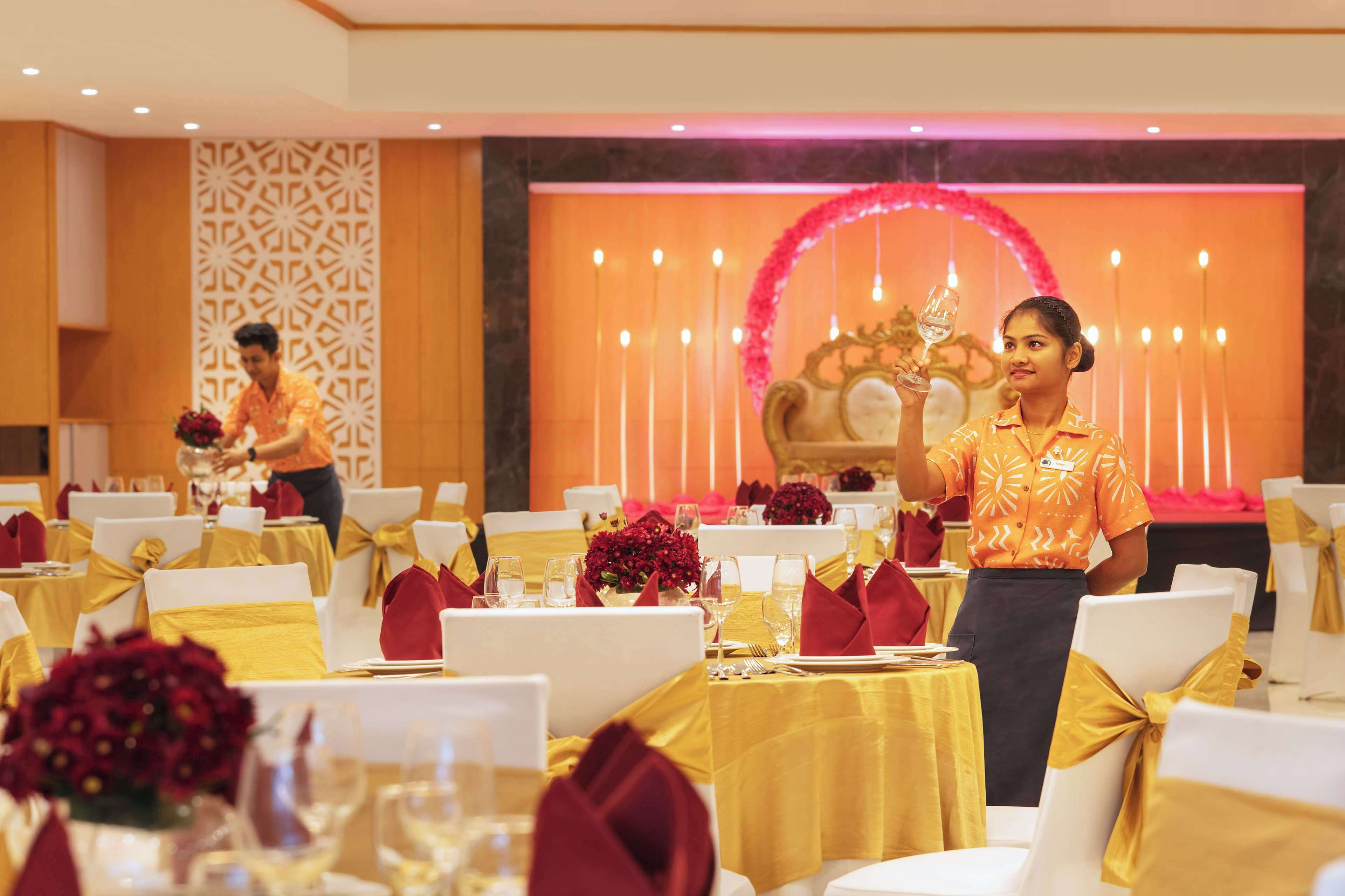 A banquet hall with tables set for a formal event, featuring white chairs with gold sashes, and a decorative backdrop with candles and flowers.