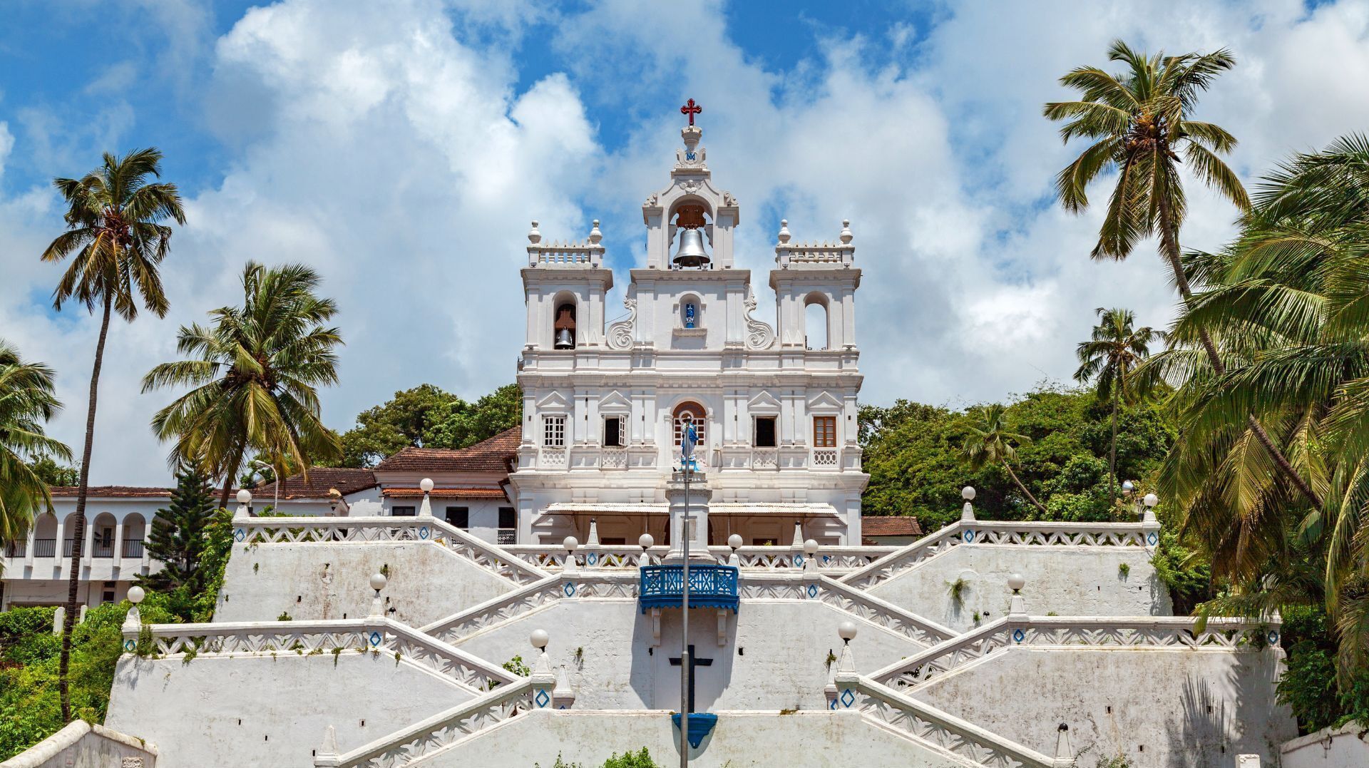A white colonial-style church with a bell tower, surrounded by palm trees and a staircase leading up to it.