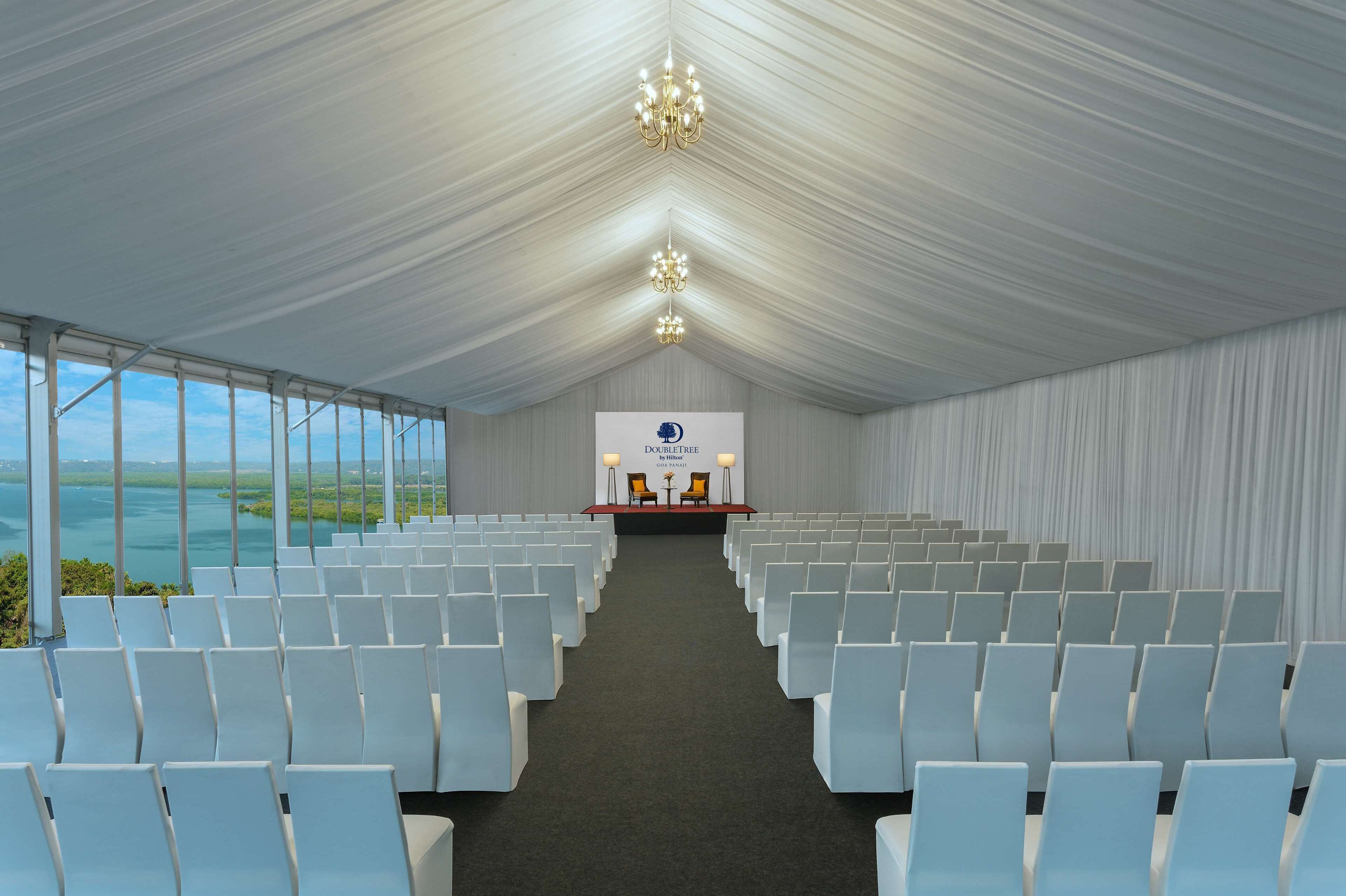 A conference room with rows of white chairs, a stage with a podium, and a view of the ocean through large windows.