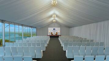 A conference room with rows of white chairs, a stage with a podium, and a view of the ocean through large windows.