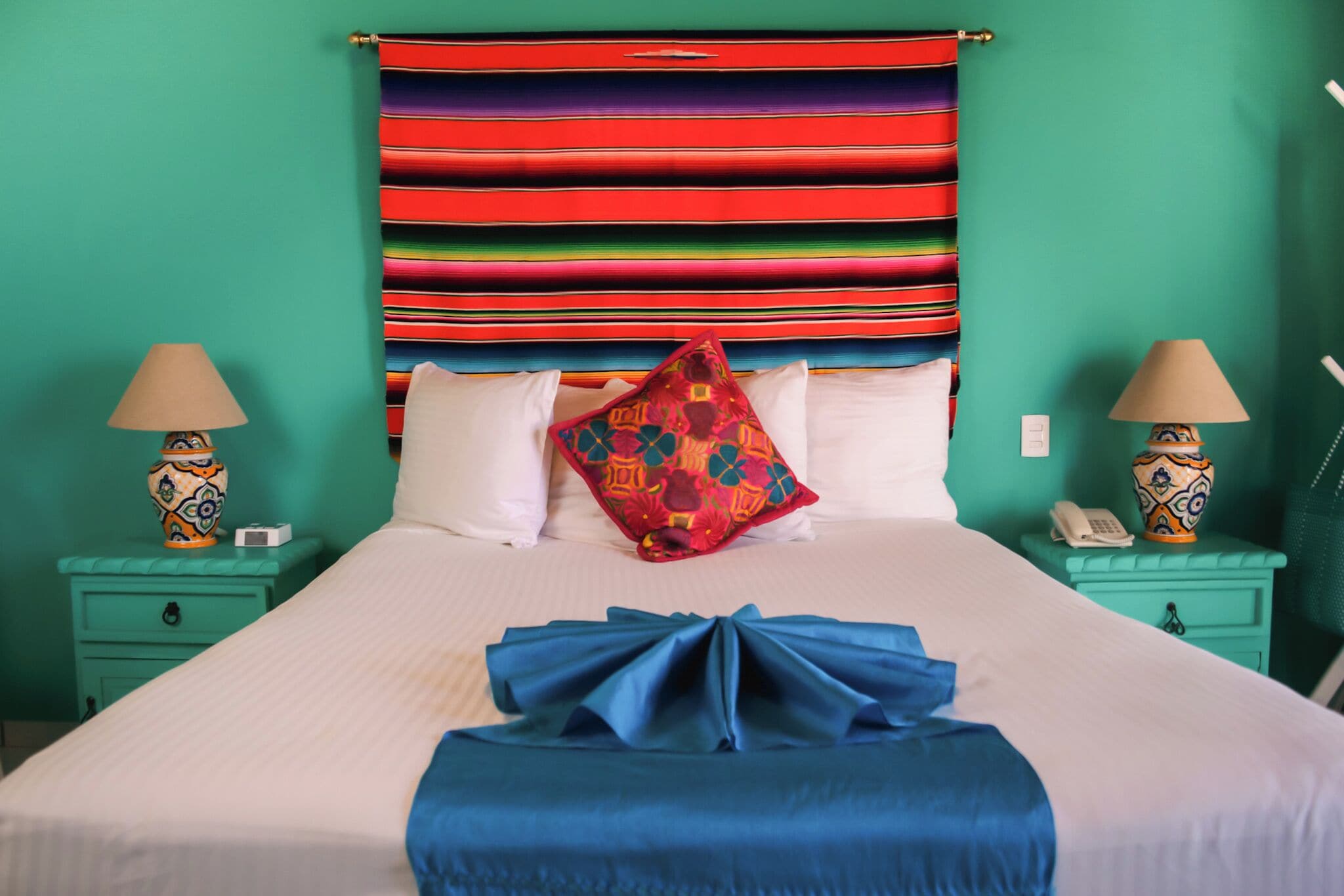 Un lit bien fait, agrémenté d’une tête de lit rayée aux couleurs vives, deux tables de chevet avec des lampes et un mur vert.