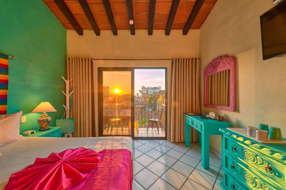 A hotel room with a bed, a desk, a dresser, and a balcony with a view of the city.
